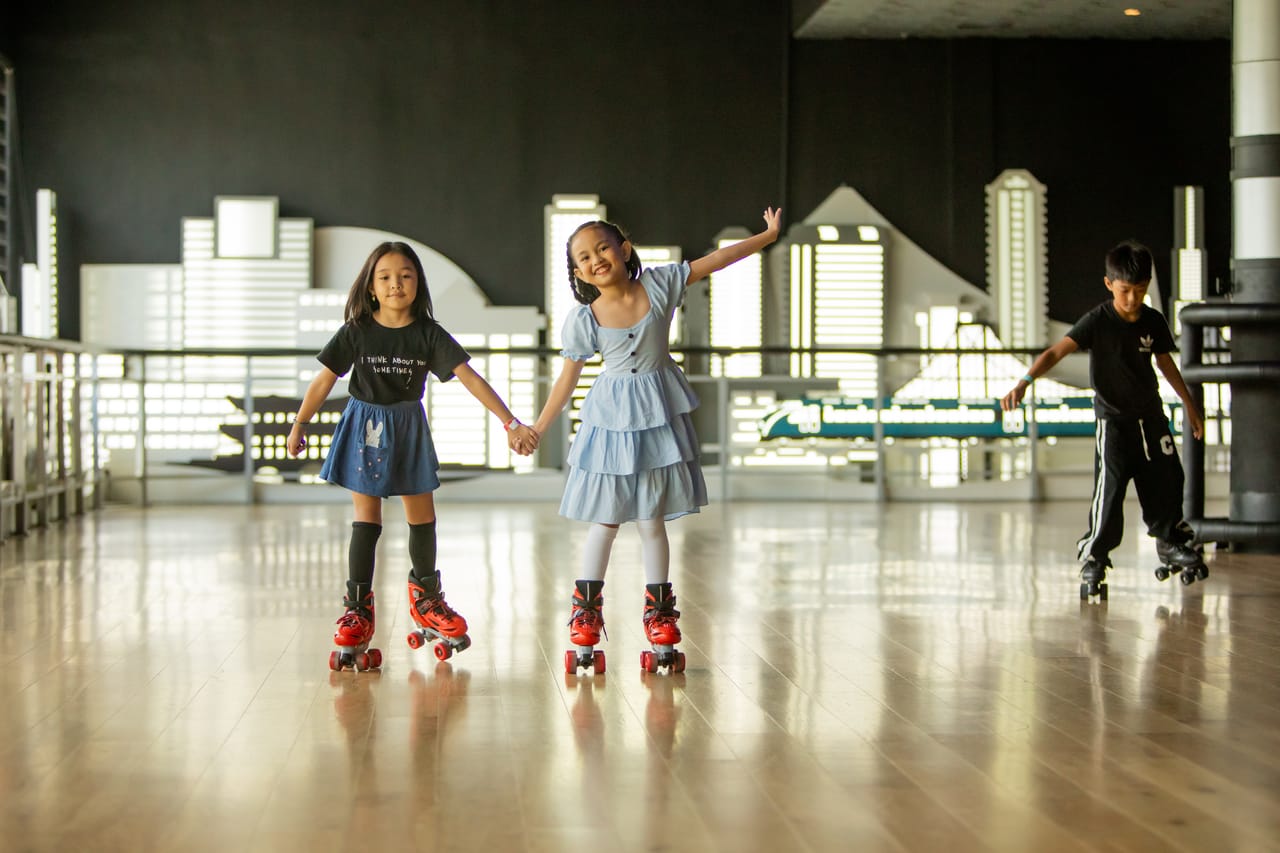 Two young girls holding hands on red quad skates at Spin City, the lit city-skyline wall glowing behind them